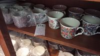 Shelf displaying multiple ceramic cups with colorful folk floral patterns and matching saucers wrapped in bubble wrap underneath