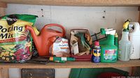 Shelf with complete gardening supplies lot including potting mix, watering can, gloves, hand cultivator, hose nozzle, Windex cleaner, and spray bottle.
