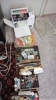 White Stella Elna sewing machine with fold out flat work surface extension on carpet. Multiple boxes and trays of sewing supplies including threads, needles, and pins arranged in front.