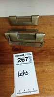 Photo showing two vintage brass lock components placed on a wooden surface, one above the other, with a MaxSold lot tag labeled 'Locks'.
