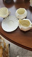 Three pieces of Belleek China set displayed on a wooden table: creamer, sugar bowl with lid, and saucer.