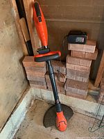 Full view of Black & Decker GrassHog 18V cordless string trimmer leaning against brick wall showing orange and black body with front handle and protective shield.