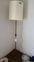 Full view of the vintage floor lamp showing metal base, slender metal pole, and off-white cylindrical cloth shade against a plain wall.