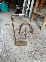 Full view of iron garden ornament with vertical stand and hanging horseshoes, showing curled decorative details and rectangular base on concrete floor.