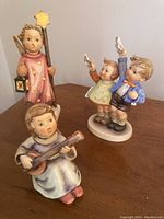 Three porcelain Hummel figurines on a wooden surface: one angel holding a lantern with a star wand, one angel playing a stringed instrument sitting down, and two children standing, each holding a bird.