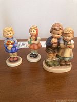 Front view of four Hummel figurines including two separate figures of a boy with a flower and a girl with a doll, and a paired figurine of a boy and a girl.