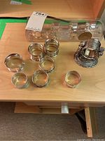 Photo showing all napkin rings arranged on a wood surface, including clear acrylic rectangular pieces in original box, several silver or silver-plated rings, and the ornate cherub napkin ring holder.