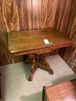 Photo showing the rectangular wooden table with carved pedestal base and caster wheels against a wood-paneled wall.