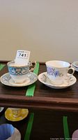 Two vintage tea cups with matching saucers, one turquoise with intricate floral design and gold trim, and the other white with purple and blue floral accents and gold trim.