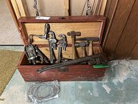 Wide view of open wooden plumber's tool box with various plumbing hand tools inside including pipe wrenches, hammers, and drill