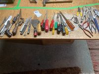 Top view of assorted hand tools on wooden surface, includes various screwdrivers with colored handles, scrapers with wooden handles, metal awl, small knives, and a few pliers.
