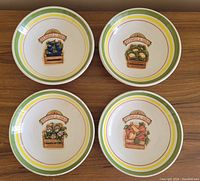 Top-down photo showing four medium/large round ceramic serving bowls with vegetable crate artwork and green, yellow, red rim bands.