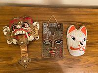 Three masks lined up on wood surface: large colorful Barong mask, rectangular carved face mask wall art, white Japanese fox mask
