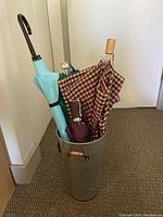 Four assorted umbrellas of varying colors and handle styles inside a tall hammered metal umbrella bin on carpeted floor against wall.