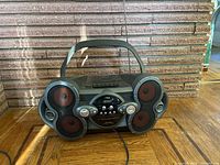 Front view of Aiwa CSD-XD55 stereo system showing dual speaker configuration with red cones, play control buttons, cassette slot, top CD player, and carrying handle.