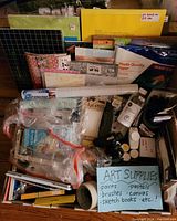Full view of the collection showing various art supplies including brushes, paint tubes, canvases, sketchbooks, pads of photo quality paper, glue sticks, masking tape, and packaging with unopened items.