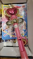 Photo of pink Cardcaptor Sakura wand with red heart top and wings, missing battery cover, placed on top of a colorful coloring book with anime character on cover.