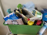 Green plastic bin filled with assorted cleaning supplies including spray bottles, sponges, microfiber cloths, and cleaning liquids