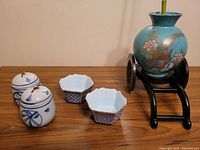 Photo of two white ceramic covered pots with blue ribbon pattern, two small hexagonal bowls, and turquoise vase with floral/bird design on black wheeled stand with bamboo.
