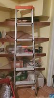Front view of 6 foot aluminum step ladder with orange plastic top and four steps, standing in front of shelves.