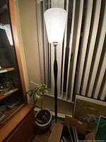 Photo of a tall floor lamp beside a window with vertical blinds. The lamp has a white conical fabric shade and a black slender stand with a subtle curve. A plant and furniture are also visible nearby.