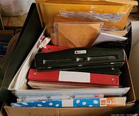 Box containing black and red binders, black 2-hole punch, assorted envelopes and file folders.