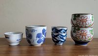 Four ceramic sake or tea cups arranged side by side on a white surface. Two have blue floral and brush stroke patterns; two have red flower and green leaf patterns stacked.