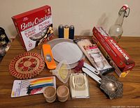 Photo showing assortment of kitchen items including Betty Crocker burner liners, mandolin slicer, colorful snack tray, wooden salt and pepper shakers, metal tea strainer, foil and wrap boxes, tongs, and other small tools.