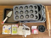 Several baking trays, pans, egg beaters with attachments, cookie cutters, rolling pin, kitchen timers, icing writer, and spatula arranged on a surface.