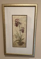 Photo of framed artwork featuring two orchid-like flowers in muted tones with textured beige background, gold-toned frame under glass.