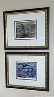 Two framed prints in black and gold frames with white matting, hung vertically one above the other on a wall, showing stylized landscape scenes.