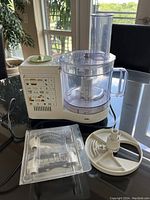 White Braun food processor base with control panel, clear plastic work bowl, power cord visible, plus attachments including slicing/shredding discs and a white plastic pusher.