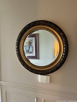 Photo shows round wall mirror hung on a light-colored wall. Frame is brass colored with a dark brown outer rim featuring pattern details.