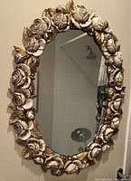 Oval mirror with three-dimensional floral frame featuring roses and leaves painted in gold and white tones.
