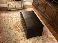 Angled top and side view of dark brown faux leather ottoman on a patterned area rug in a living room, showing its rectangular shape and padded stitched top.