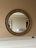 Front view of the round wall mirror hanging on a beige wall above a white mantle, showing the detailed brass-colored frame with leaf-like patterns.