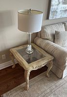 Wooden side table with glass panel top placed beside a sofa, featuring carved legs and edges. A lamp with a white shade is on top of the table, but the lamp is not included in the lot.