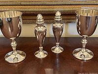 Two silver-plated goblets and two salt and pepper shakers on wooden surface in front of a framed mirror.