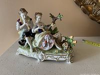 View of the front and side of the porcelain figurine showing the seated couple, musical instruments, dog, floral details, tree, and ornate base with a measuring tape for scale.