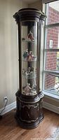 Full view of rounded vintage wood and glass curio cabinet, lighted interior, situated near window.