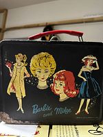 Front view of black metal lunchbox with colorful Barbie and Midge illustrations, shows rust and wear, red handle visible