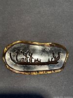 Front view of brooch showing raised silver design of boat and figures within a bronze border with bear paw stamps.