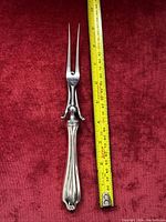 Full length photo of the carving fork with a yellow measuring tape beside it showing about 10.5 inches length on a red fabric background.