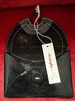 Photo of new black leather wallet with white stitching, circular flap and button snap closure with attached tag.