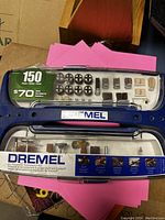 Photo of the closed blue hard plastic case of Dremel accessory kit featuring label indicating 150 pieces and $70 retail value. Visible sanding drums, grinding stones, polishing wheels, cutting bits.