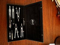 Open black case displaying six silver metal drawing compasses and accessories arranged in foam slots, branded Kern Swiss inside lid