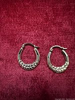 Photo of two small hoop earrings made of 10k gold with detailed leaf pattern design, displayed on red fabric background