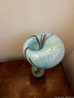 Photo showing the top and partial side of a blue glass sculptural form with gold swirls, flared curled top, on a wooden surface against a beige wall.