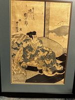 Framed Japanese woodblock print showing a man seated in traditional robes inside an interior setting, with Japanese calligraphy and stamps visible.