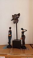 Three figurines displayed together: two African women figures and one abstract winged figure on a black pedestal.